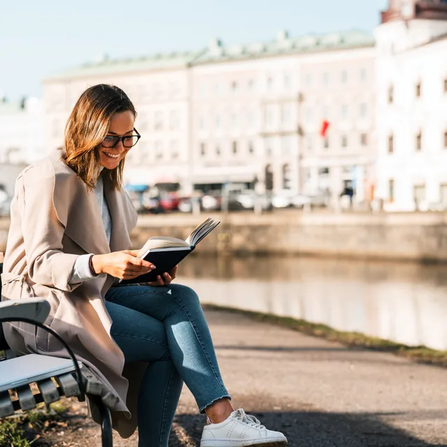 A woman sitting on a bench reading a book outdoors, representing a relaxed way to learn advanced German while enjoying a scenic environment.

