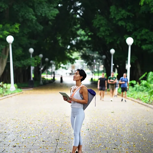 Woman holding a tablet while walking through a tree-lined park, representing mobile learning for online French classes.