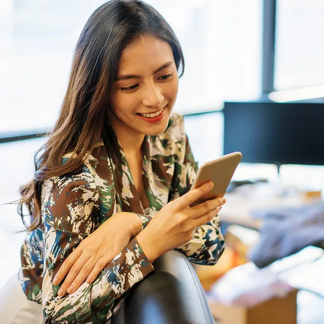 A woman in an office setting, smiling while using her phone, representing a modern approach to global corporate language training.