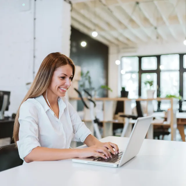 Smiling young woman with a laptop at a modern workspace, exploring opportunities in a French teaching career.