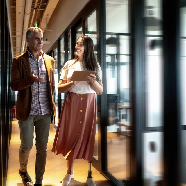 Two colleagues in a modern office hallway discussing strategies for a German teaching career, with a young woman holding a tablet and wearing a prosthetic leg.