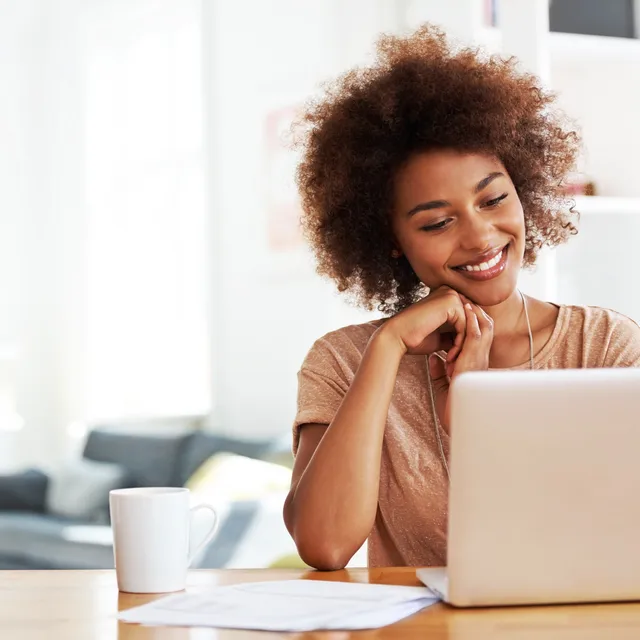 Woman smiling at her laptop while attending Italian classes online from a cozy home setup with a mug and papers on the desk.