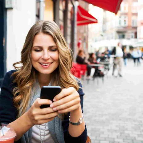 Woman smiling while using her smartphone at a café, representing the convenience of Berlitz online language schools for mobile learners.
