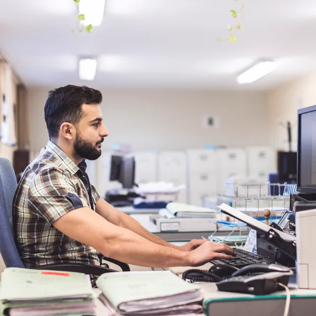 Man taking a written English test on a desktop computer in a quiet office setting, focused on typing.

