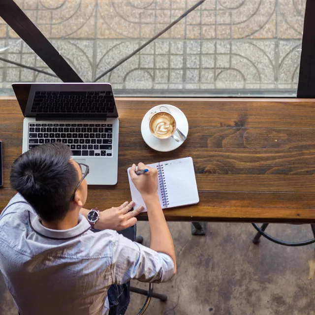 Student taking notes at a café table while attending online Portuguese classes on a laptop, illustrating the flexibility of Berlitz language programs.

